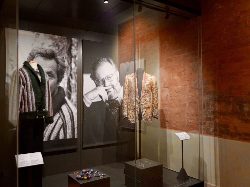 Display case at Palazzo Morando’s “The Gentleman” exhibition in Milan with two menswear jackets, men’s jewelry on pedestals, and large black-and-white portrait backdrops.