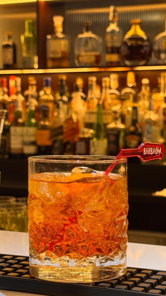Close-up of the Rabo de Galo cocktail served in a crystal glass with ice and an orange twist garnish, sitting on a bar counter with blurred bottles in the background.