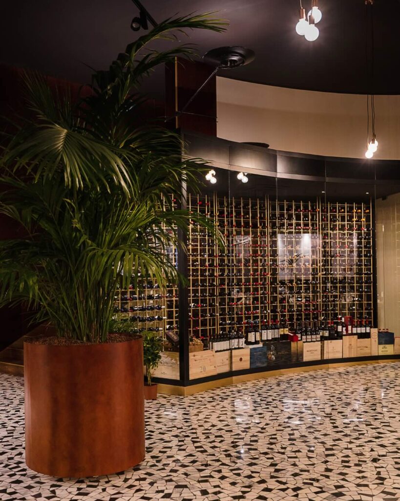 A modern, illuminated glass wine cellar displaying rows of wine bottles in the entrance of Barbacoa Milano restaurant.