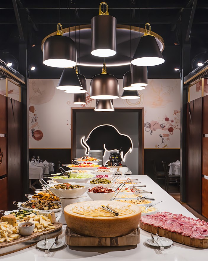 A wide view of a luxurious restaurant salad bar featuring wheels of parmesan cheese, sliced prosciutto, and various bowls of fresh salads and vegetables under modern lighting.