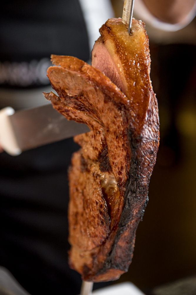 A juicy, cooked cut of beef rump steak resting on a dark slate serving board next to jars of green chimichurri sauce.