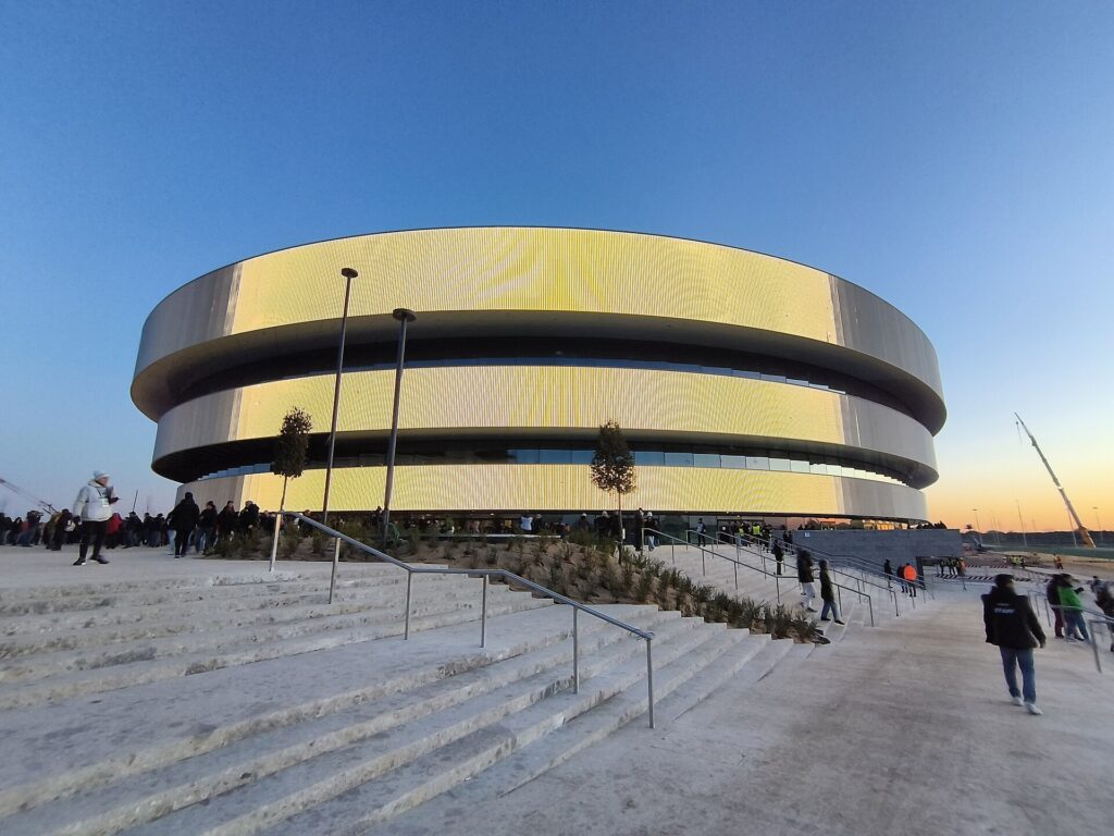 Exterior view of the Milano Santagiulia Hockey Arena at dusk. The modern, oval-shaped building features a glowing facade with illuminated yellow and gold panels. In the foreground, a wide concrete staircase leads to the plaza, with people walking around and newly planted small trees. The sky is clear blue, fading to a warm orange on the horizon.