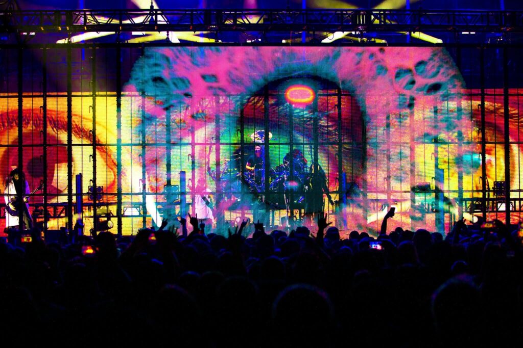 Korn live on stage in front of a large crowd, with colorful screens, dramatic lighting, and a giant eye graphic dominating the concert backdrop.